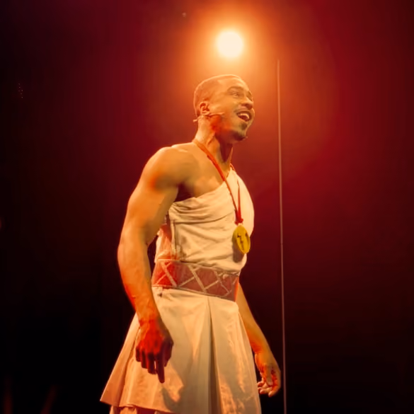 Man in a toga-like costume with a medal, singing or speaking on stage with warm lighting behind him.