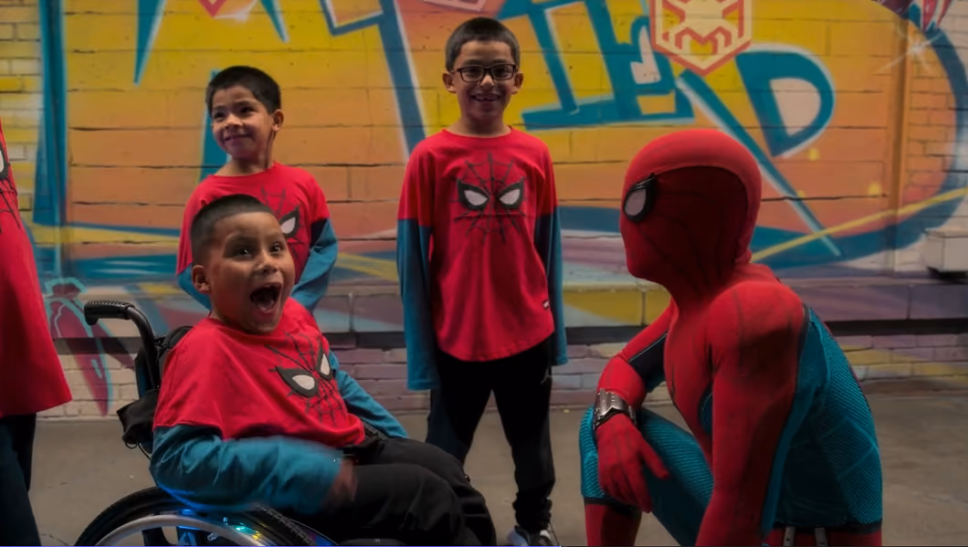 Three boys wearing Spider-Man shirts smiling near a person dressed as Spider-Man in front of graffiti wall.