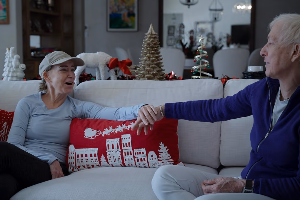 Two elderly people sitting on a white couch holding hands and smiling with Christmas decorations behind them.