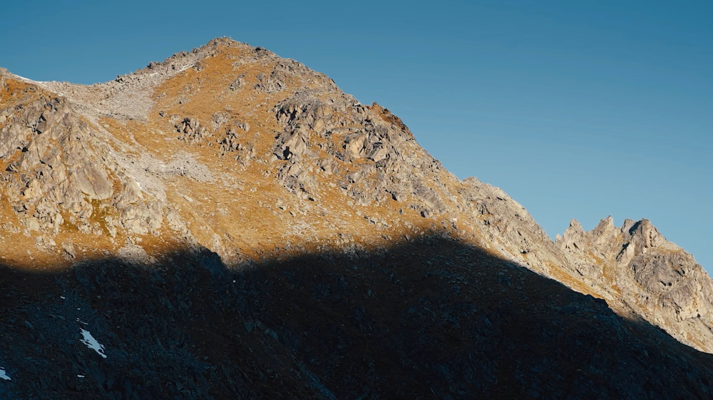 Rocky mountain peak with sunlight on upper slopes and shadow on lower slopes under blue sky.
