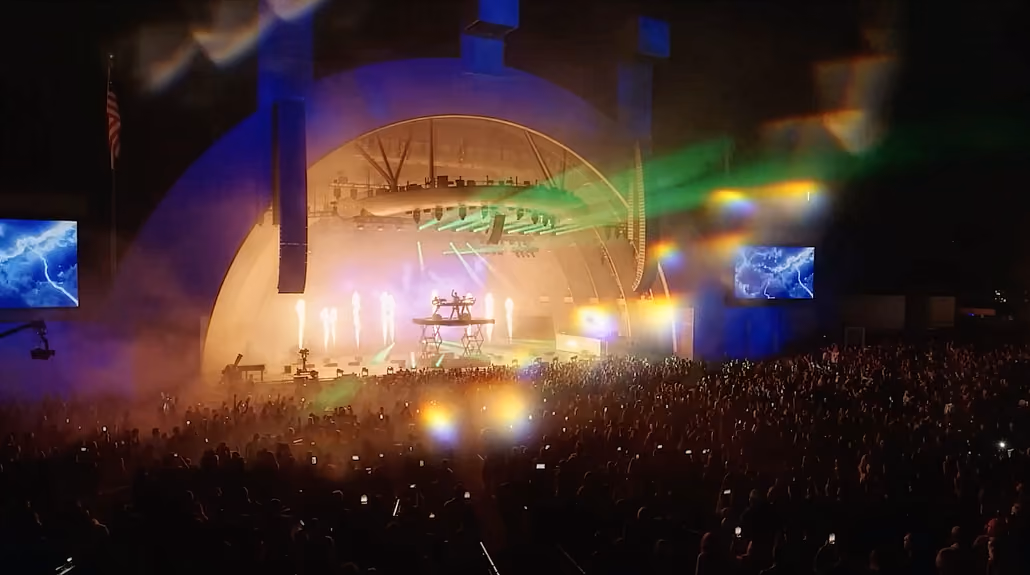 Crowd at a nighttime outdoor concert with bright stage lights and colorful lasers.