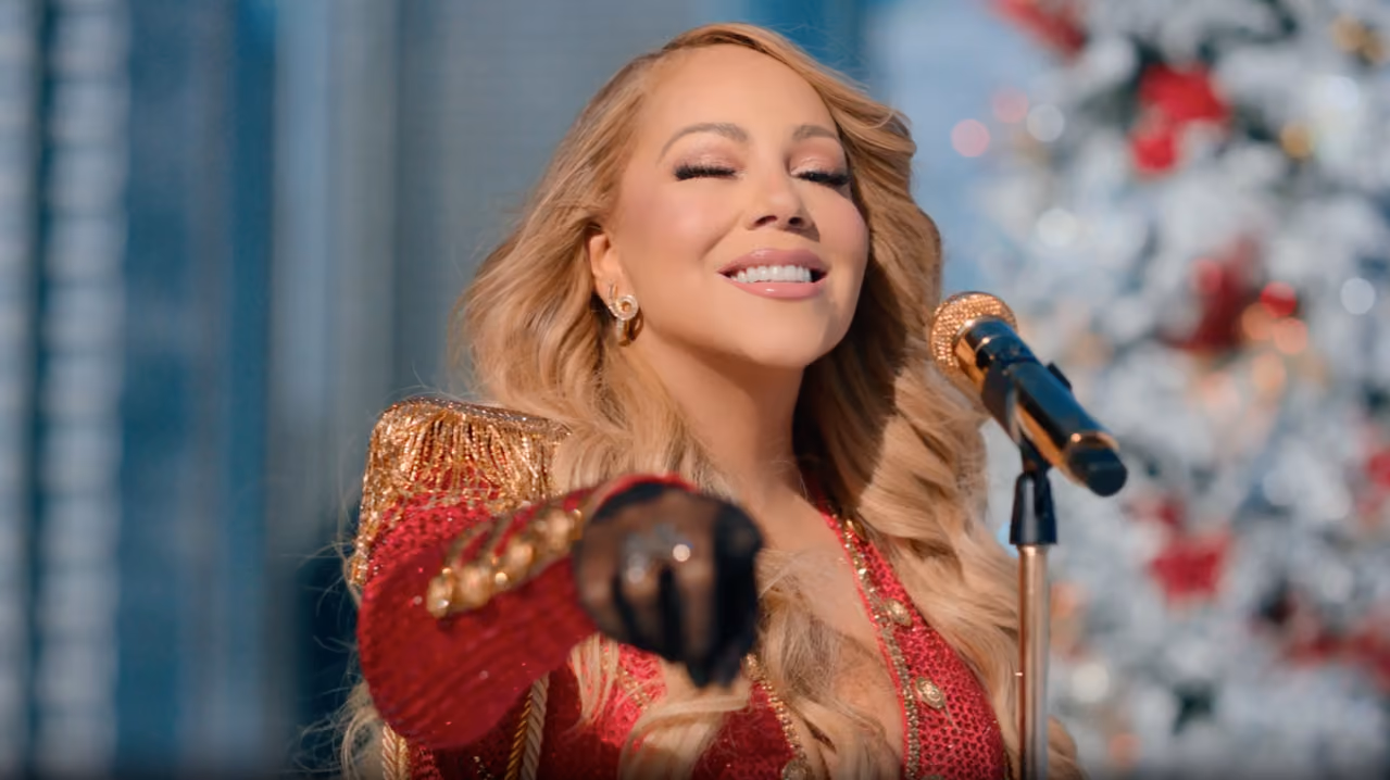 Smiling woman with long blonde hair in red and gold outfit points forward near a decorated white Christmas tree.