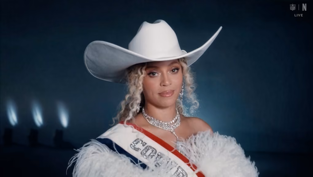 Woman wearing a white cowboy hat, sparkly necklace, sash, and feathered outfit against a dark background.