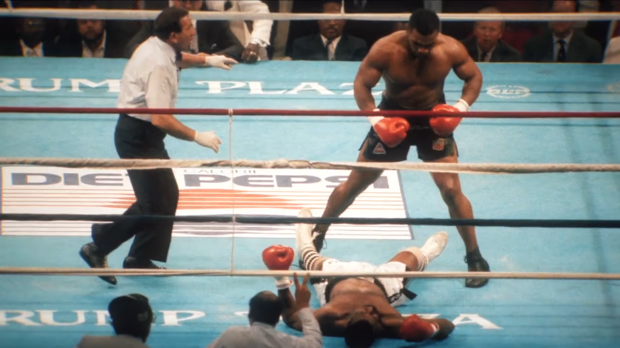 Boxer standing in ring with gloves while opponent lies on the mat and referee approaches.