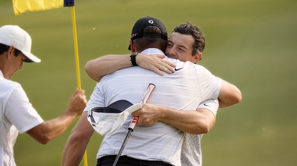 Two male golfers in white shirts embrace near a yellow flag on a golf course.
