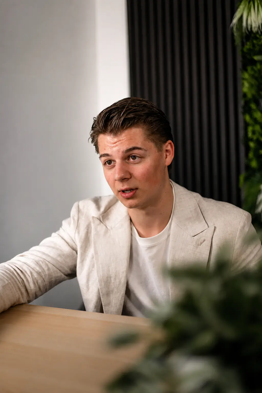 Gründer von Impression Marketing in hellbeiger Jacke spricht an einem Holztisch vor dunklem Hintergrund mit Pflanzen.
