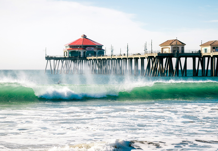 Ocean wave breaking near a wooden pier with buildings and clear sky above.