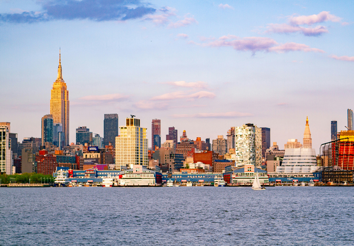 New York City skyline at sunset with Empire State Building and a sailboat on the river.