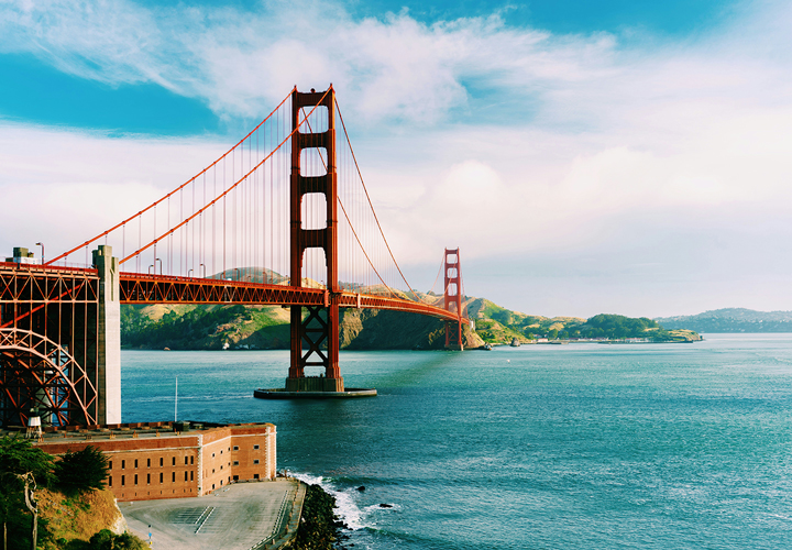 Golden Gate Bridge over blue water with green hills and partly cloudy sky in the background.