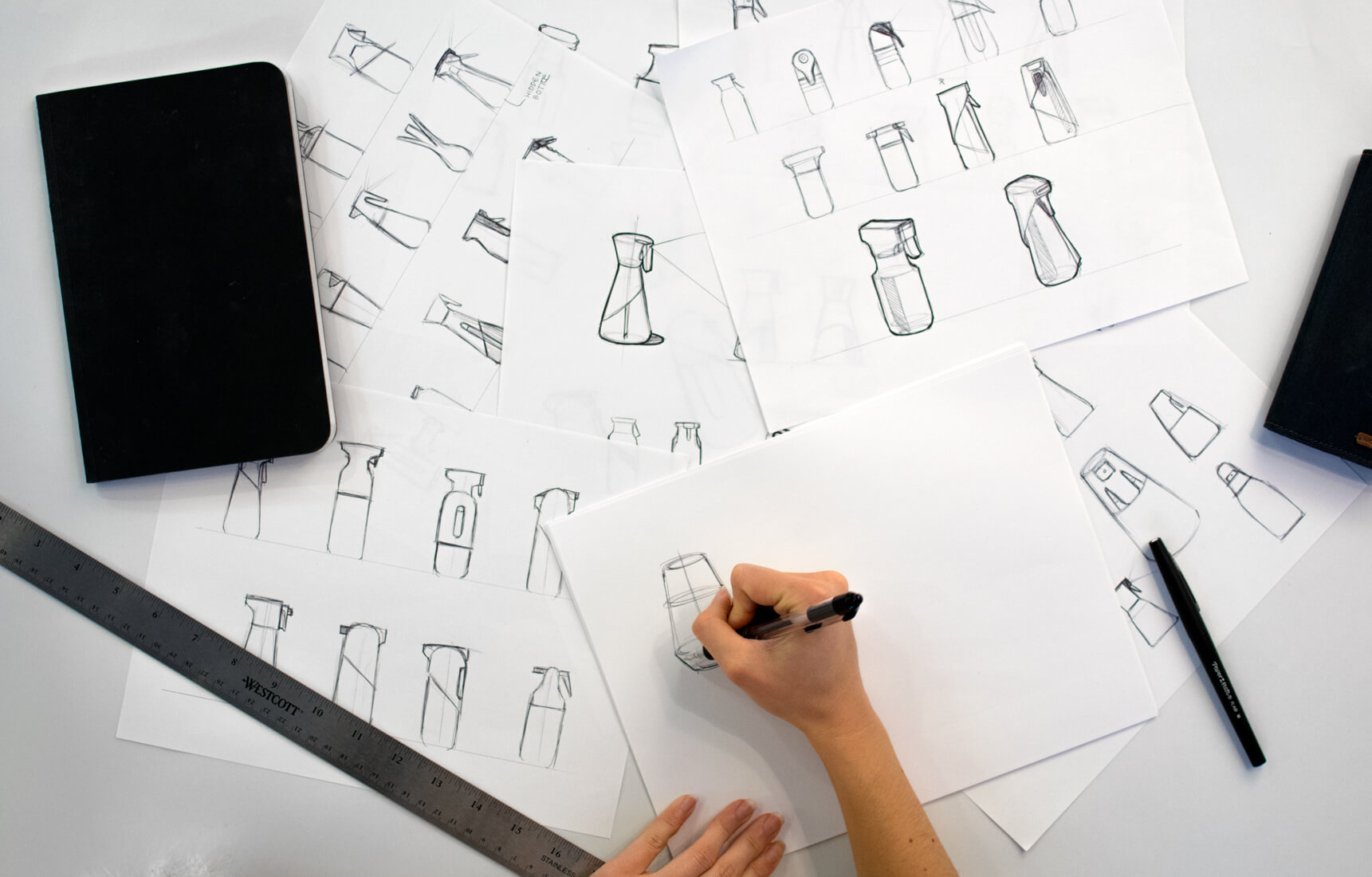 Person sketching various bottle and spray bottle designs on white sheets with a ruler and pens nearby.