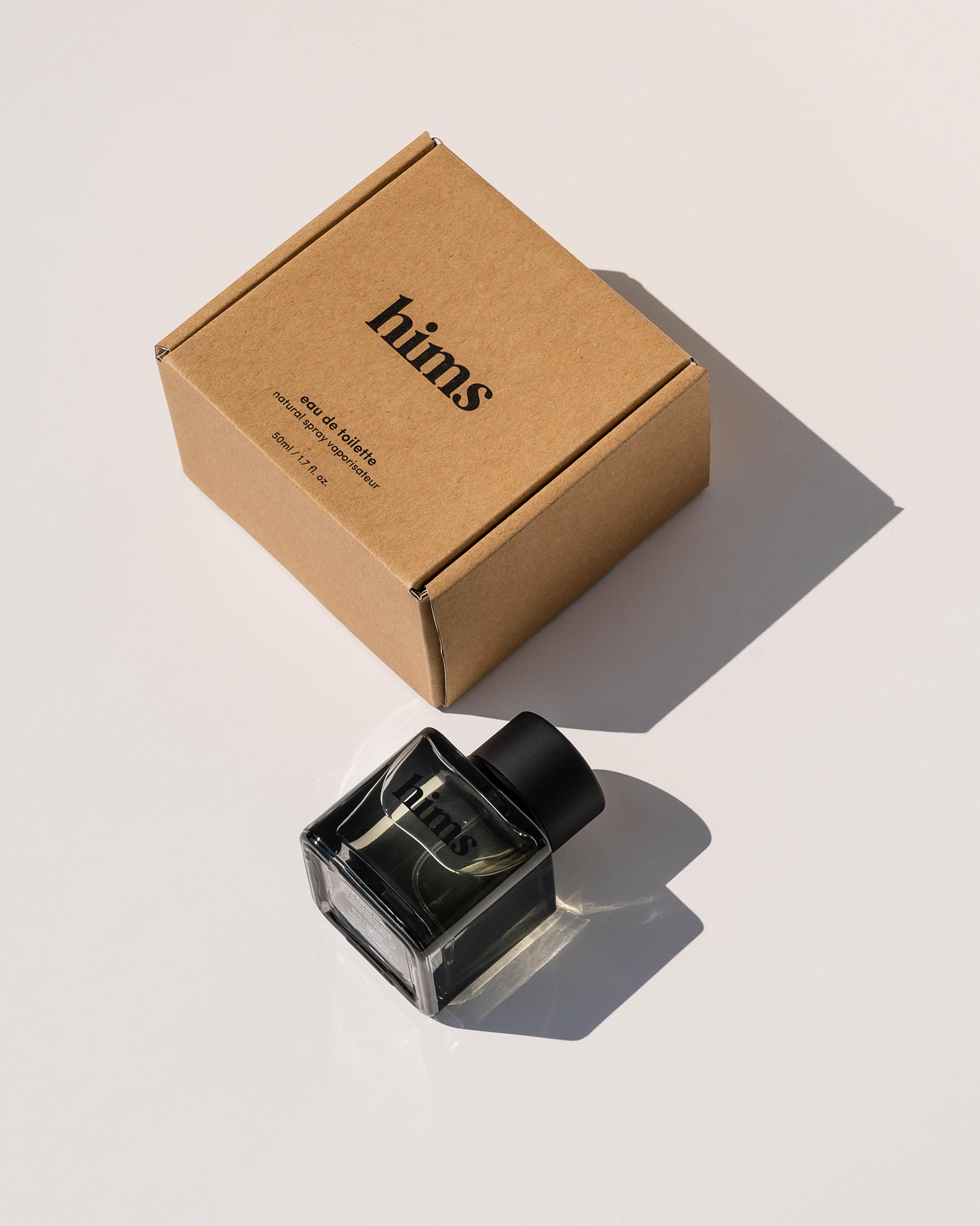 Box and black glass bottle of hims eau de toilette on white surface casting shadows.