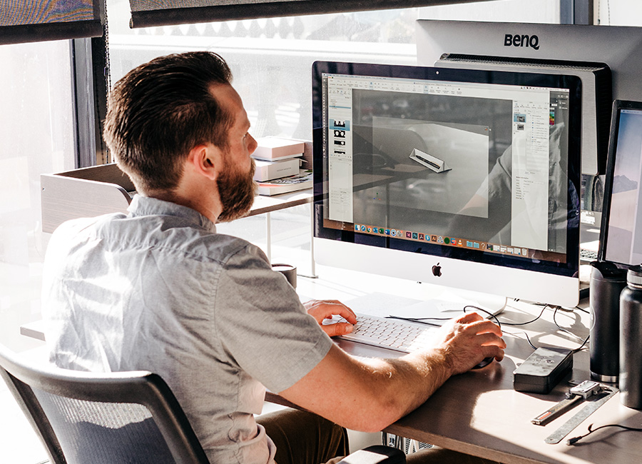 Man with beard working on 3D modeling of product packaging on an Apple computer in a bright office.