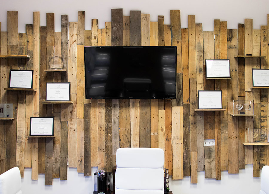 Office wall with vertical wooden planks, a large TV, framed certificates, and white chairs below.