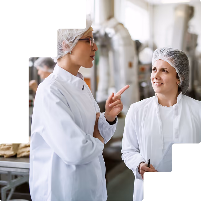 Two workers in hairnets and white coats talking on a food production floor