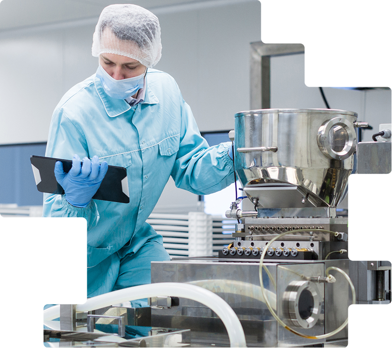 Technician in a hairnet, mask, and blue gloves reviewing production equipment with a tablet in a cleanroom