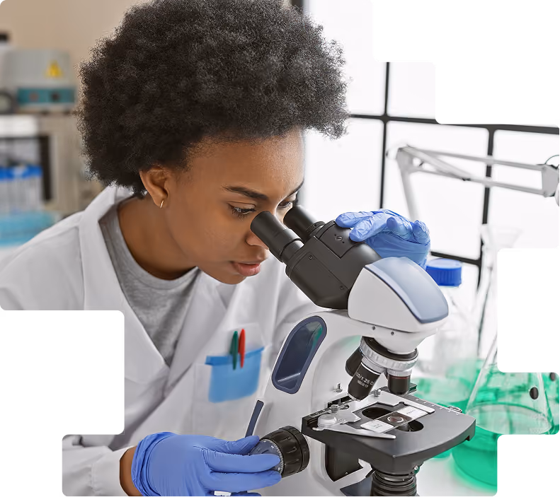 Lab technician in a white coat and blue gloves examining a sample through a microscope