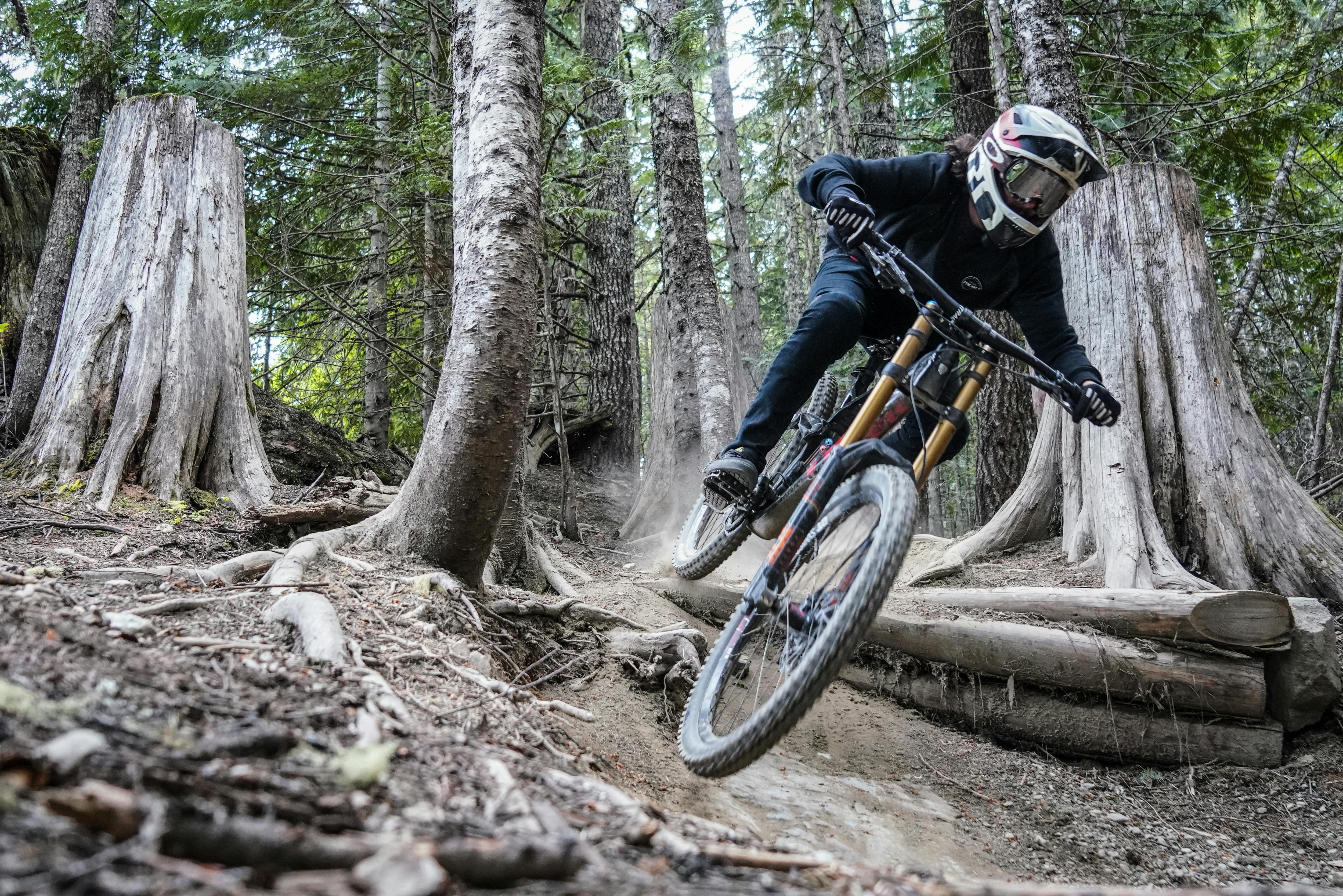 Mountain biker wearing a helmet and goggles riding downhill on a dirt trail in a forest with tree trunks and roots.