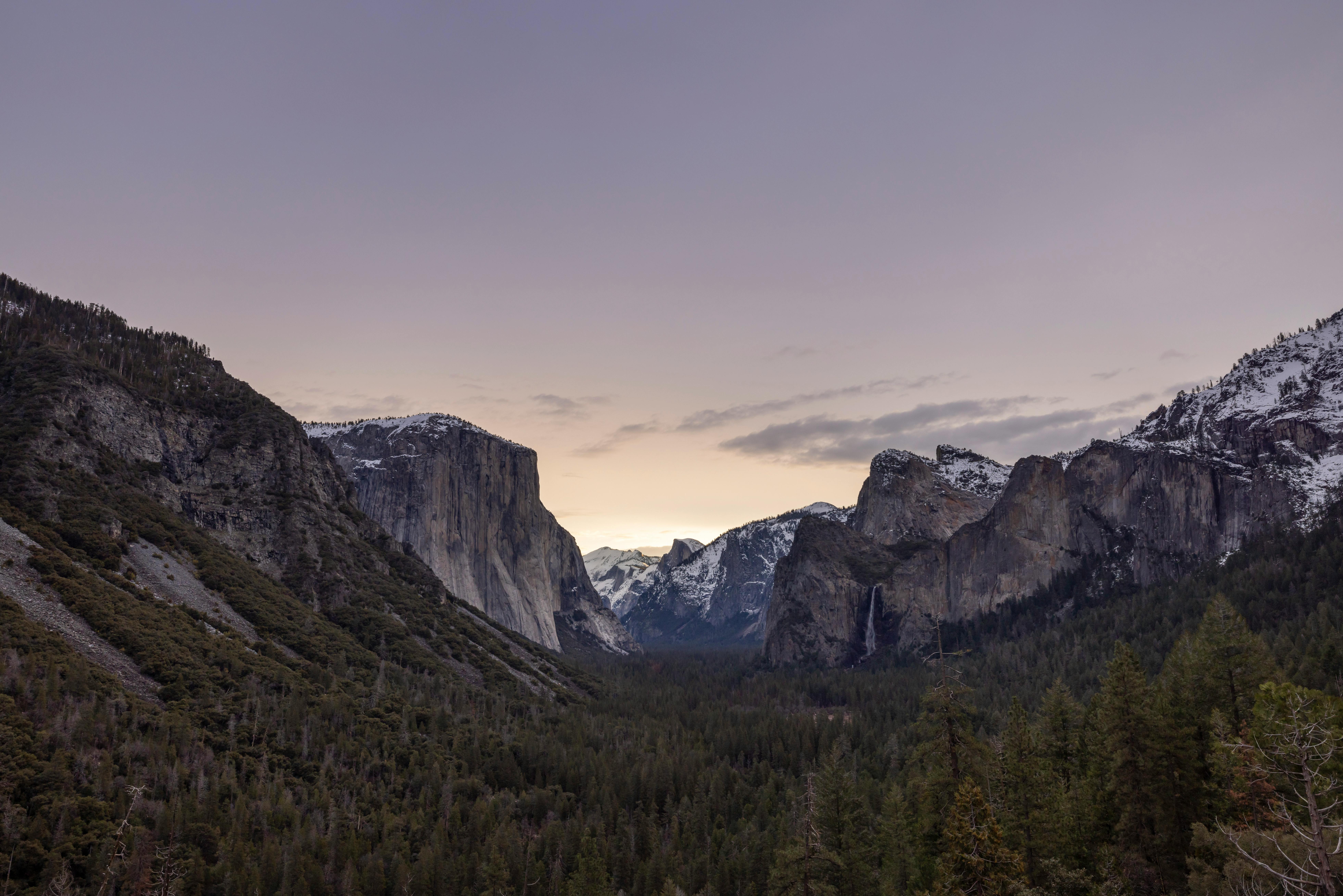 Wide valley view of Yosemite National Park with forested foreground, large granite cliffs including El Capitan on the left, and Bridalveil Fall waterfall on the right under a purple sunset sky.