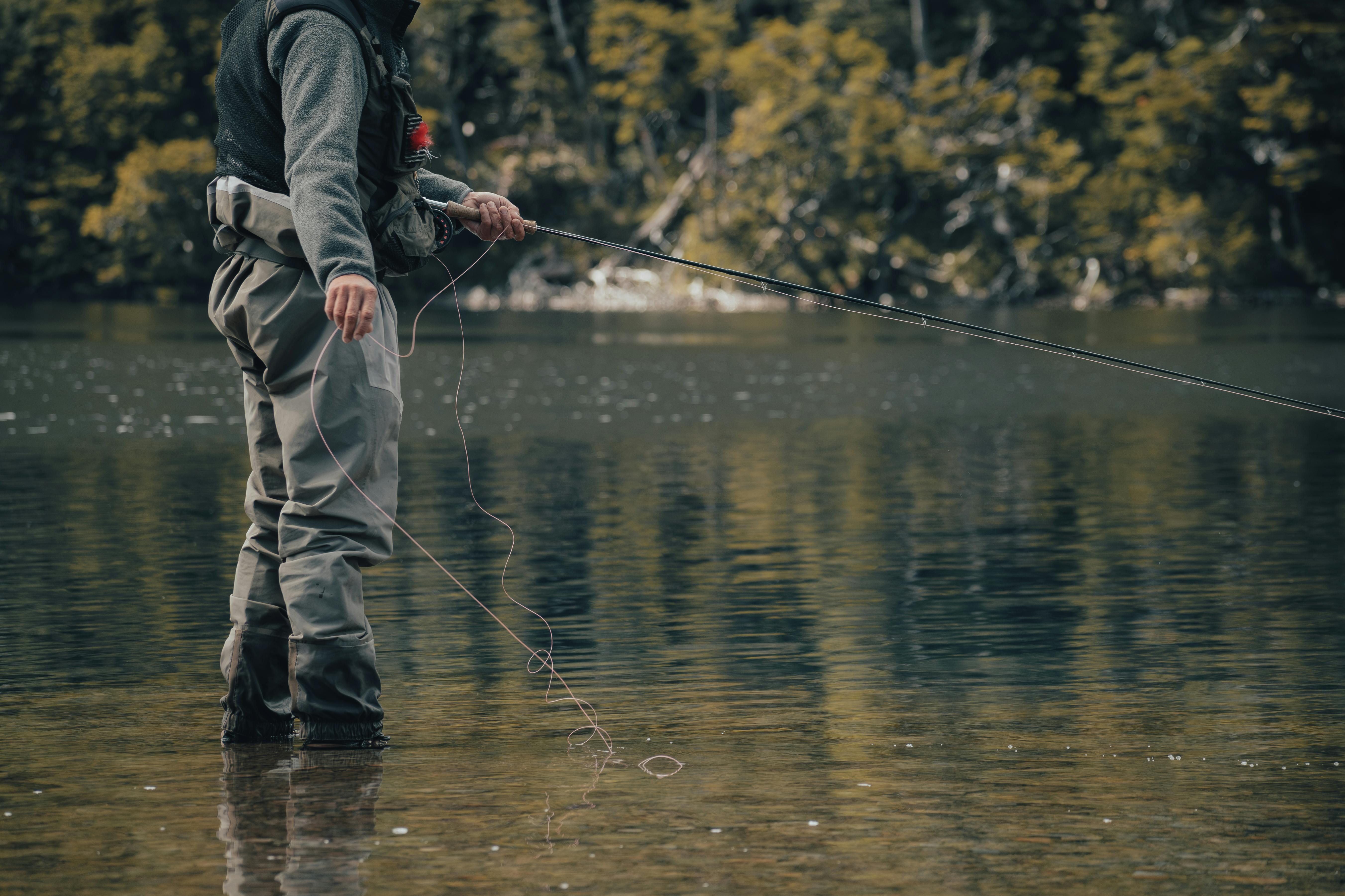 Person wearing waders fly fishing in a calm river with reflection of trees in the water.