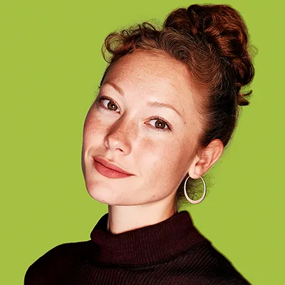 Portrait of a young woman with red curly hair in a bun, wearing hoop earrings and a dark turtleneck against a green background.