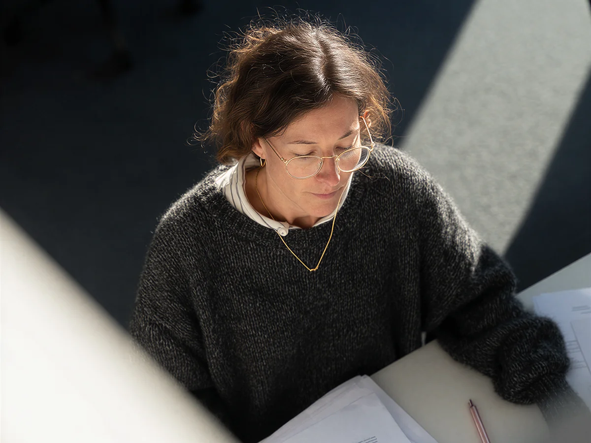 Woman wearing glasses and a dark sweater reading documents at a desk in natural light.