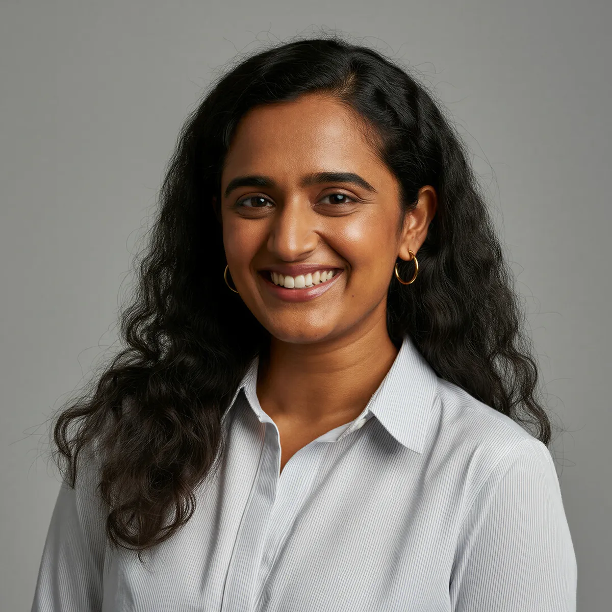 Smiling woman with long curly black hair wearing a white collared shirt and gold hoop earrings against a gray background.