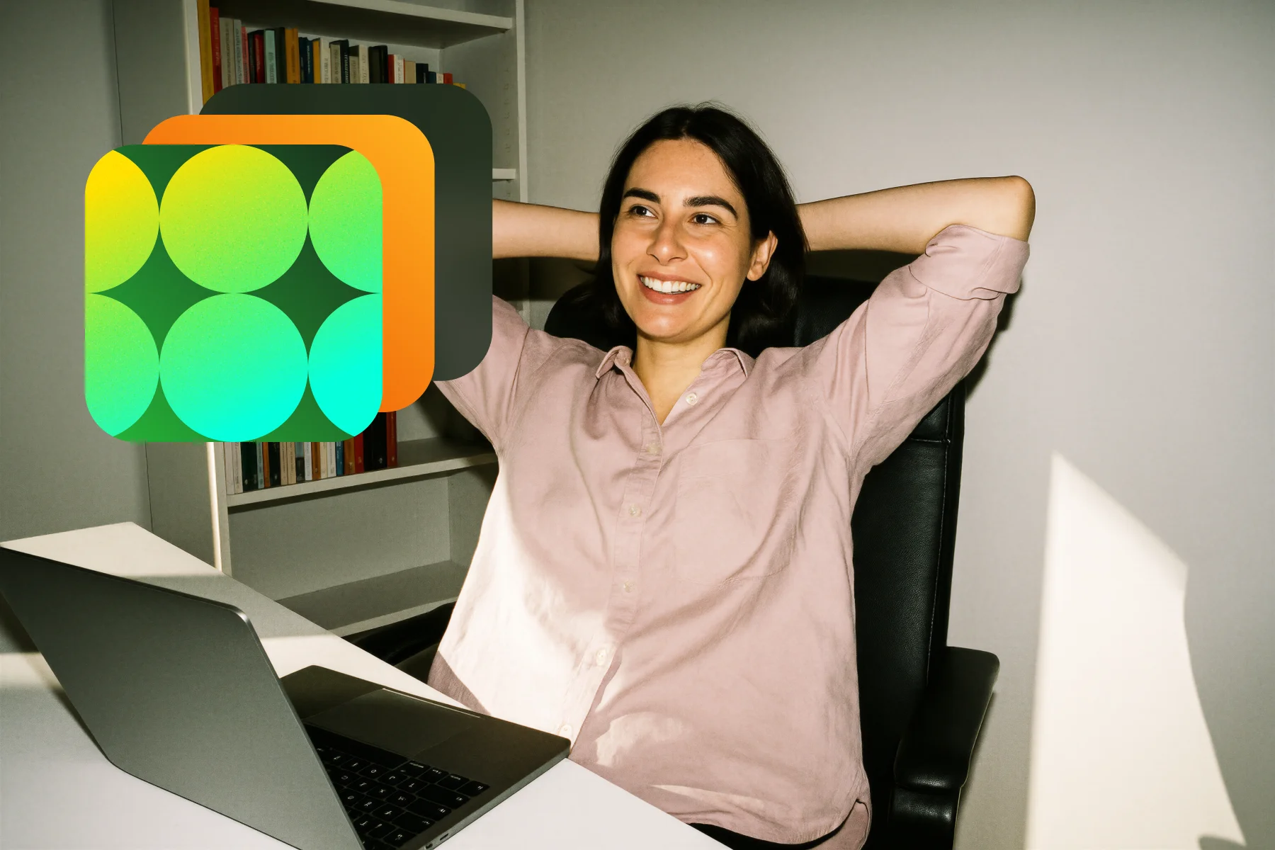 Smiling woman leaning back in chair with hands behind her head at desk with laptop, bookshelf in background.