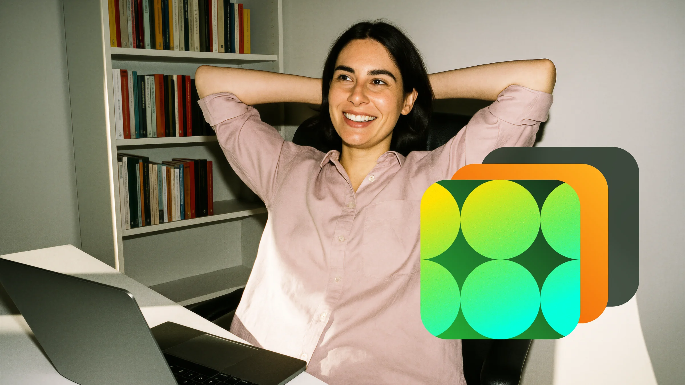 Smiling woman in a pink shirt sitting relaxed with hands behind her head by a desk with a laptop and a bookshelf in the background.
