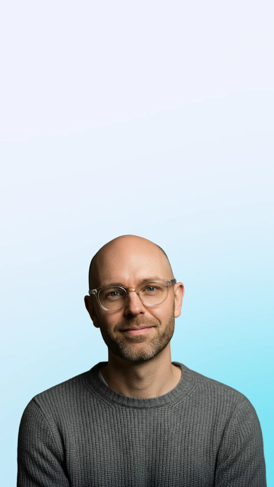 Smiling bald man with glasses wearing a gray knit sweater against a light blue gradient background.