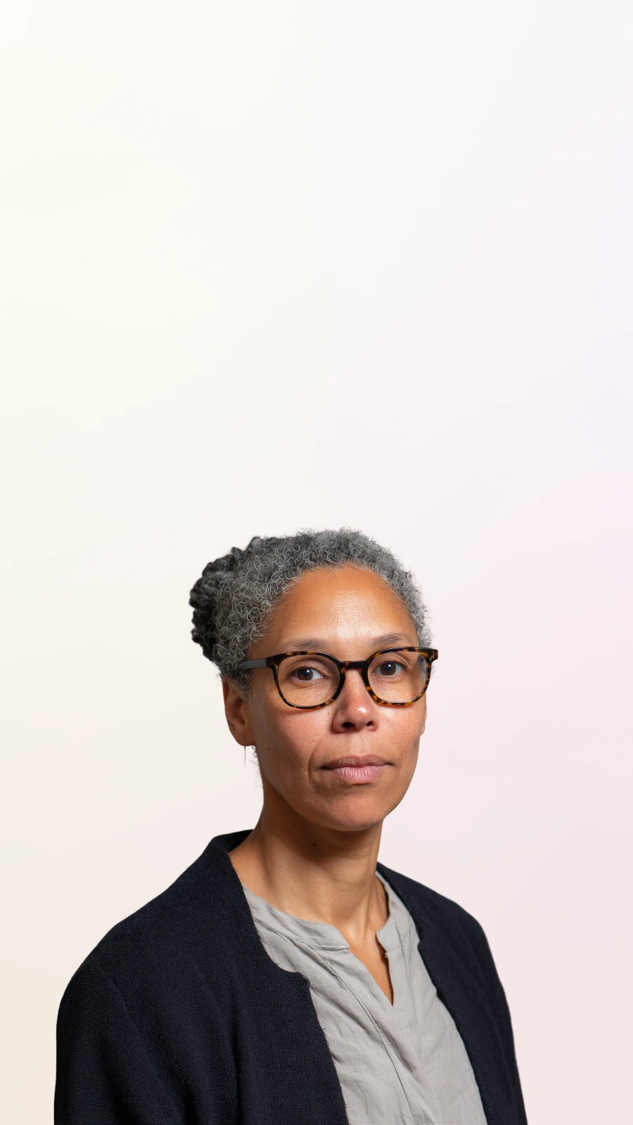 Middle-aged woman with curly gray hair in a bun, wearing tortoiseshell glasses, a gray blouse, and a dark jacket against a light background.