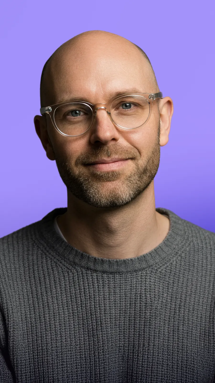 Smiling bald man with glasses wearing a gray knit sweater against a purple background.