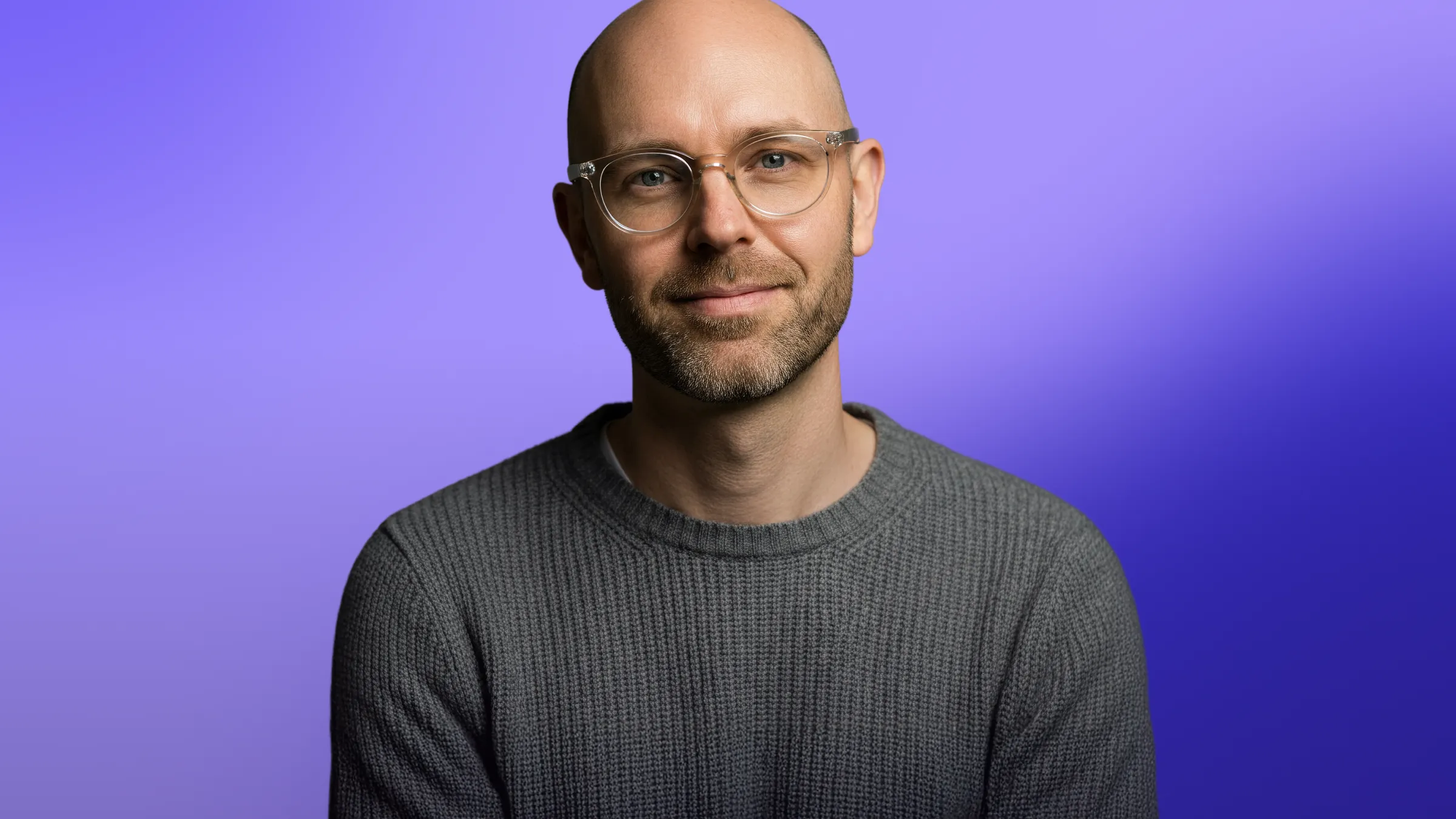 Smiling bald man with glasses wearing a gray sweater against a purple gradient background.