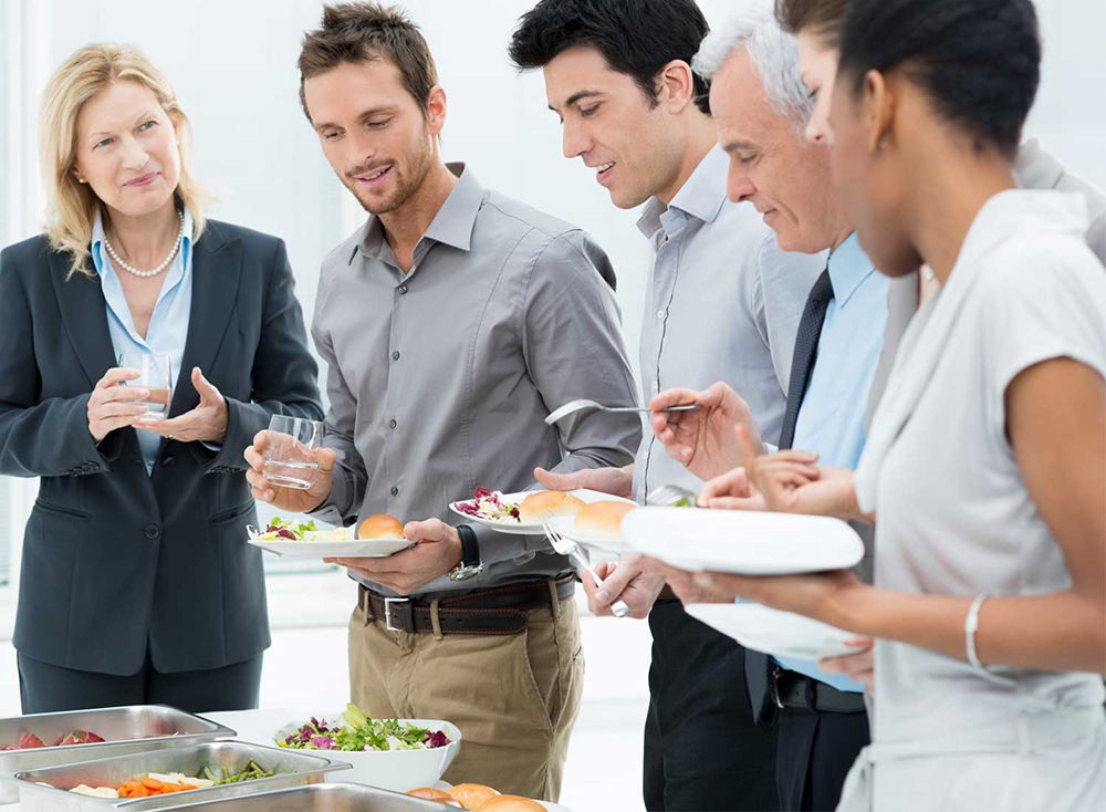 Group of five professionally dressed people standing in line at a buffet table serving themselves salad and rolls.