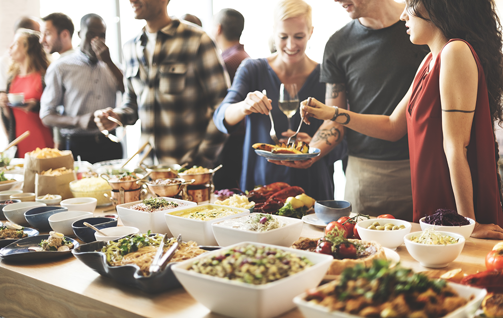 Group of diverse people serving themselves food from a buffet with various dishes on a wooden table.
