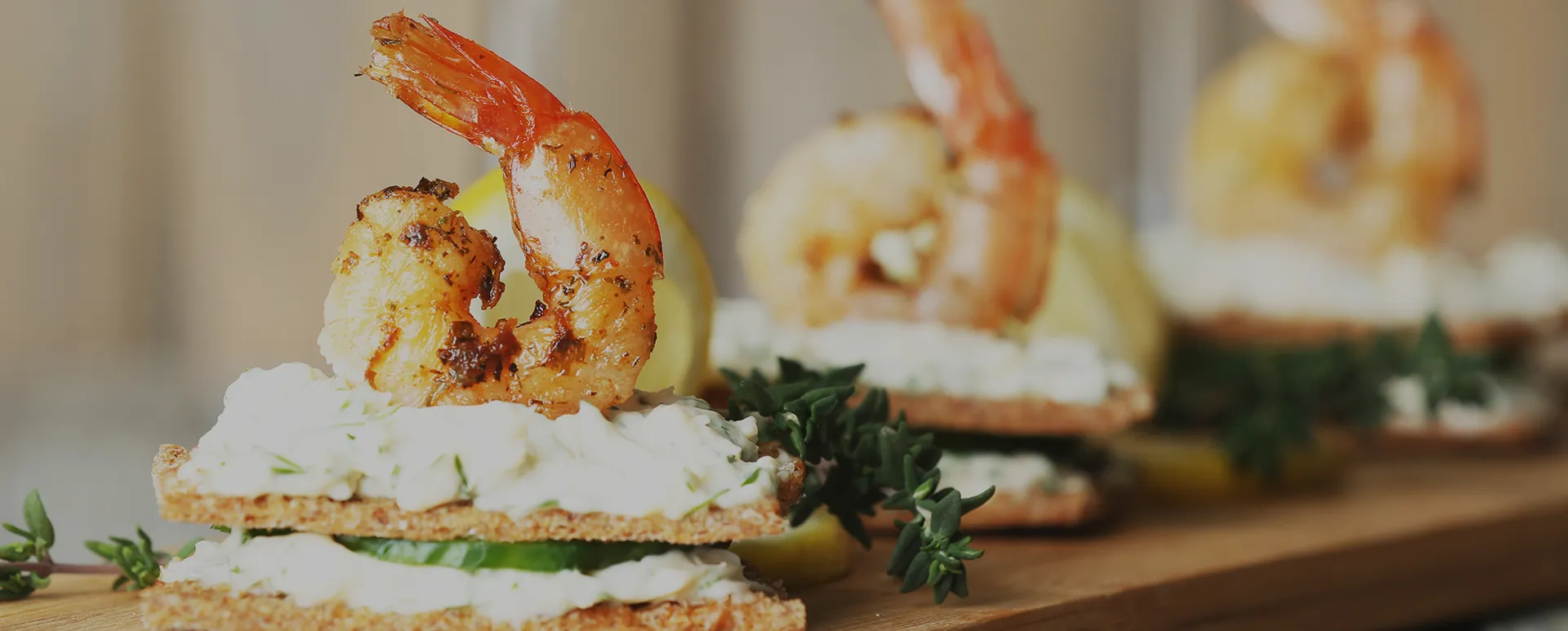Grilled shrimp served on crispbread with creamy spread, cucumber slice, and fresh herbs on a wooden board.