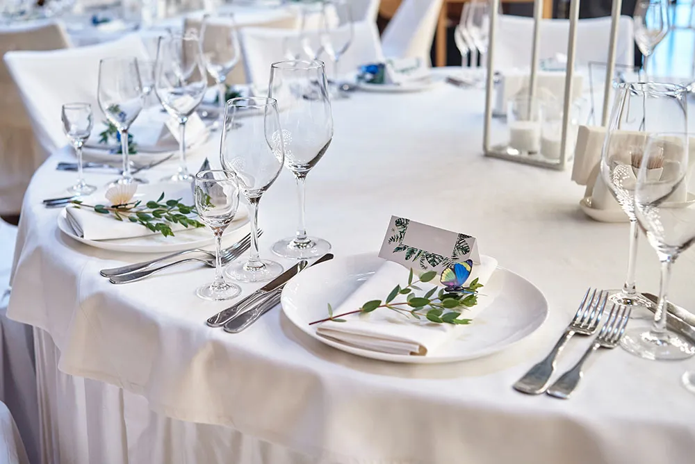 Elegantly set round table with white tablecloth, glassware, silver cutlery, white plates, folded napkins, greenery, and a name card with a blue butterfly decoration.