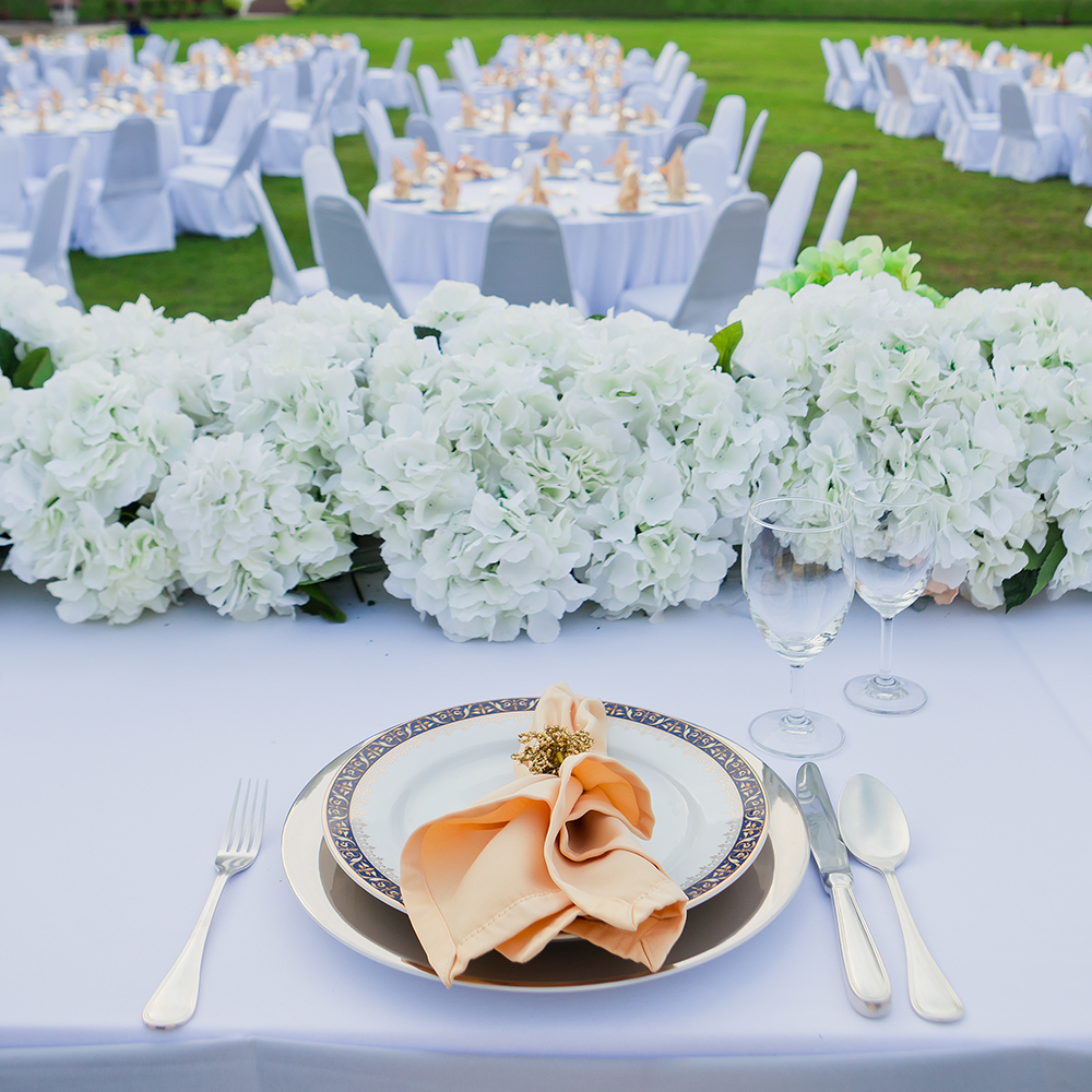 Elegant outdoor wedding table setting with white flowers, gold-rimmed plates, peach napkin, and glassware on white tablecloth.
