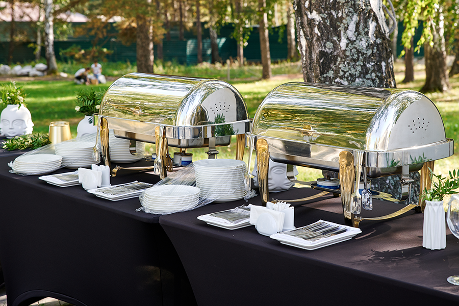 Outdoor catering table set with two closed silver chafing dishes, stacks of white plates, and neatly arranged cutlery on black tablecloths in a park setting.