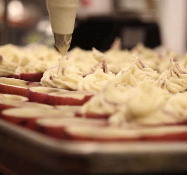 Piping mashed potatoes onto halved red-skinned potatoes on a baking tray, preparing a dish.