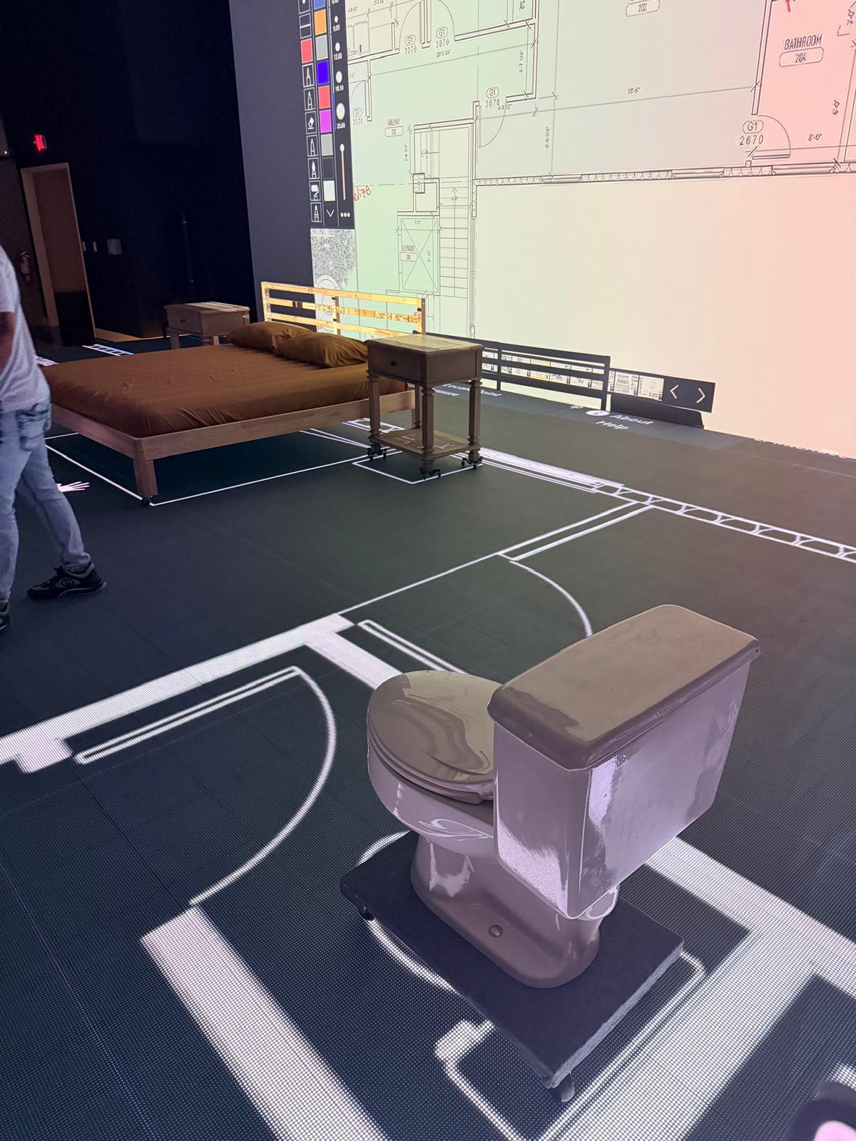 Room setup with a bed, nightstand, and toilet placed on a floor with projected architectural floor plans and a large floor plan displayed on the wall.