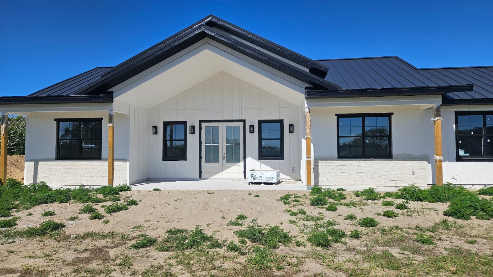 Front view of a modern white custom home with black framed windows, double front doors, and a metal roof under a clear blue sky.