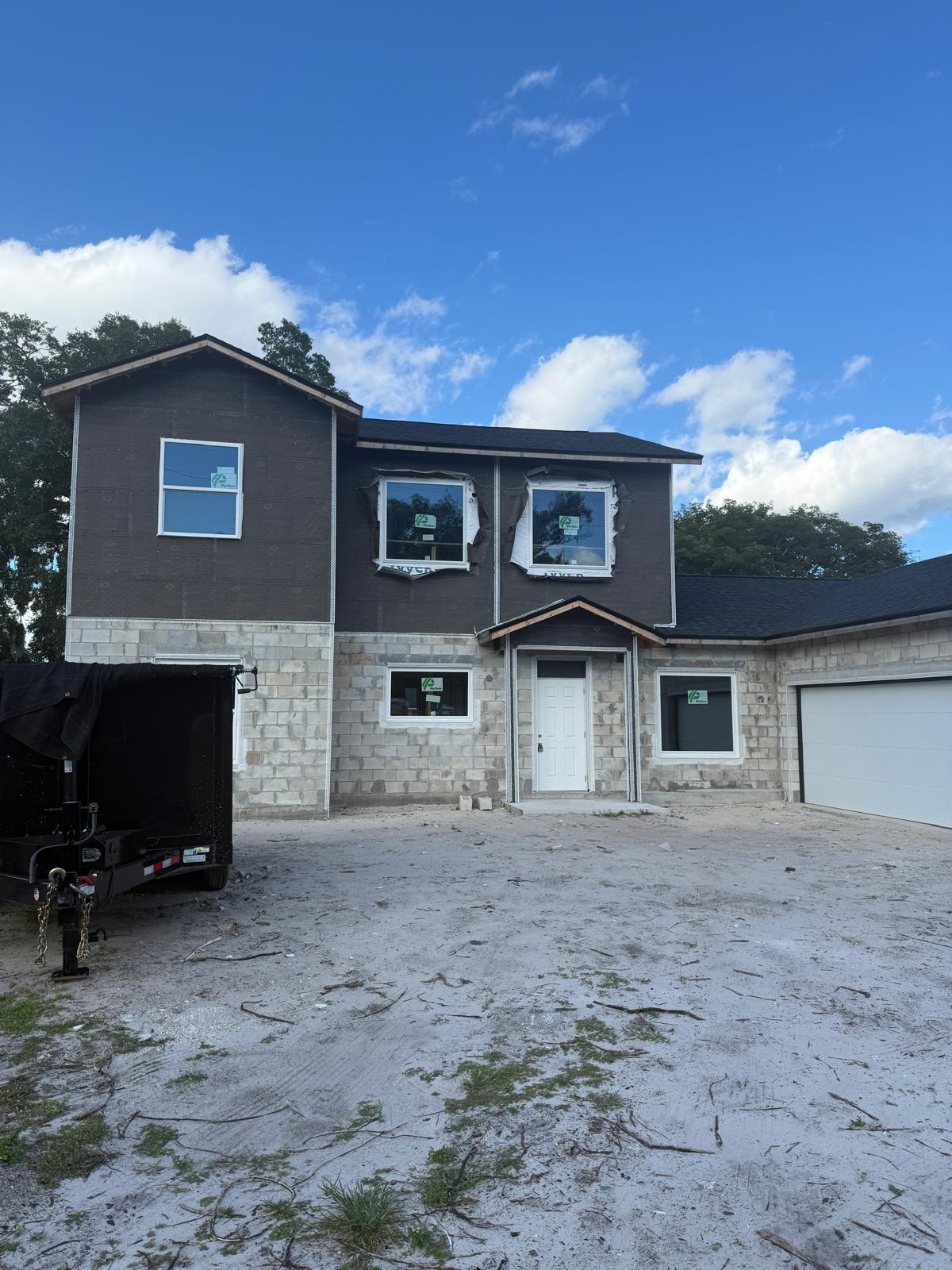 Two-story house under construction with unfinished brick walls, multiple new windows, a white front door, attached garage, and a dirt driveway.