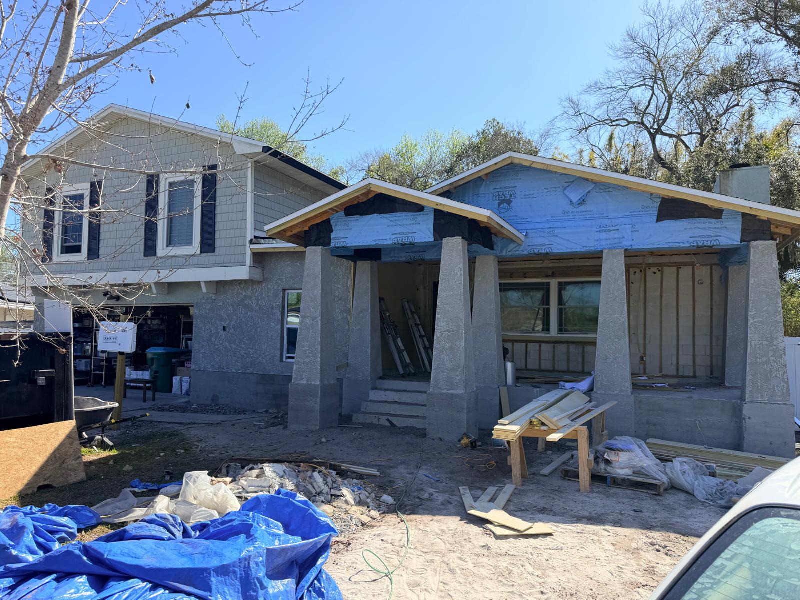 Partially constructed house with gray stucco columns and blue house wrap under a clear blue sky, surrounded by construction materials and equipment.