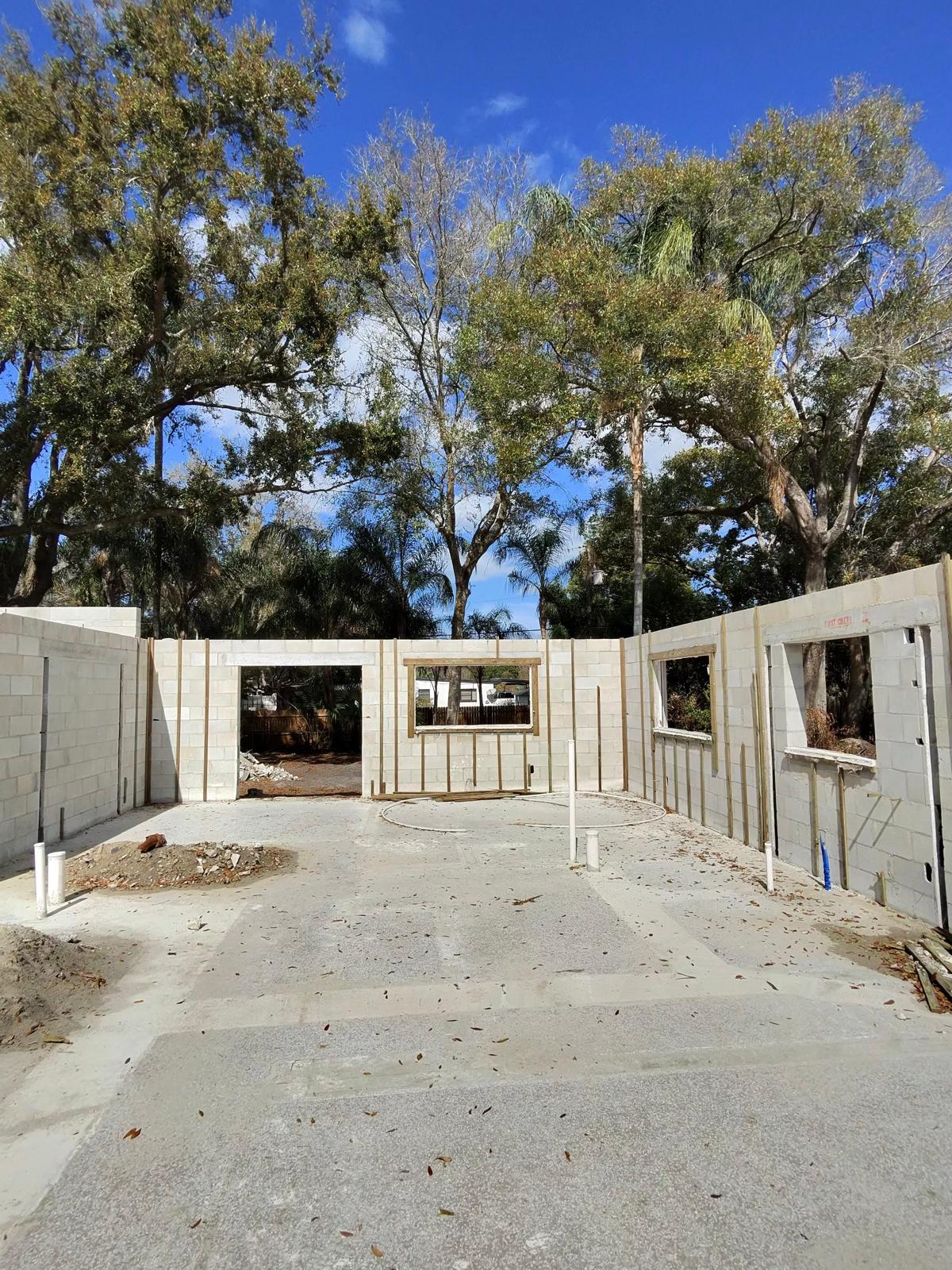 Construction site with concrete foundation and partially built block walls framing window and door openings under a blue sky with trees.