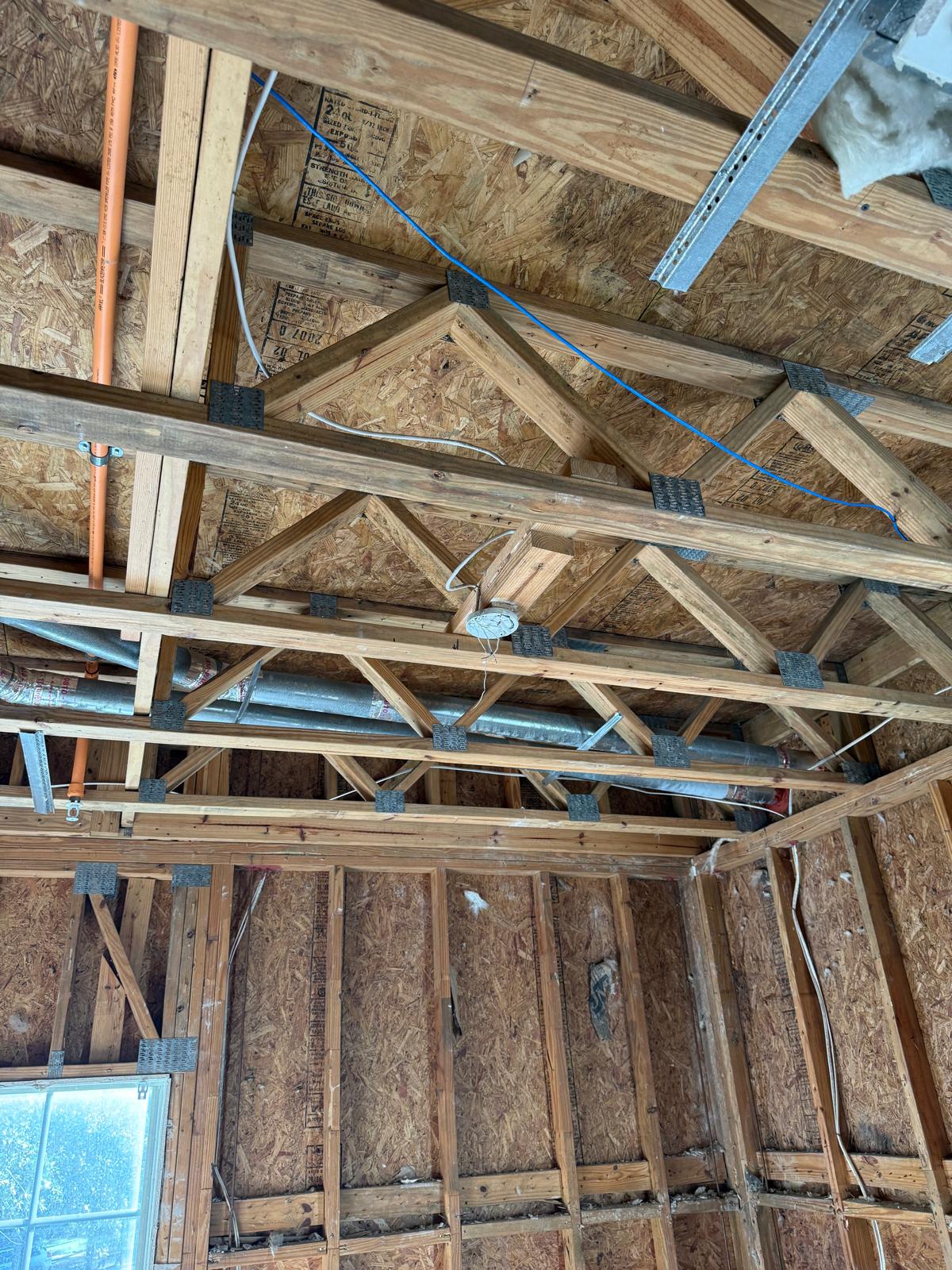 Interior view of a wooden roof frame with exposed beams, metal brackets, ductwork, wiring, and insulation materials in an unfinished building.