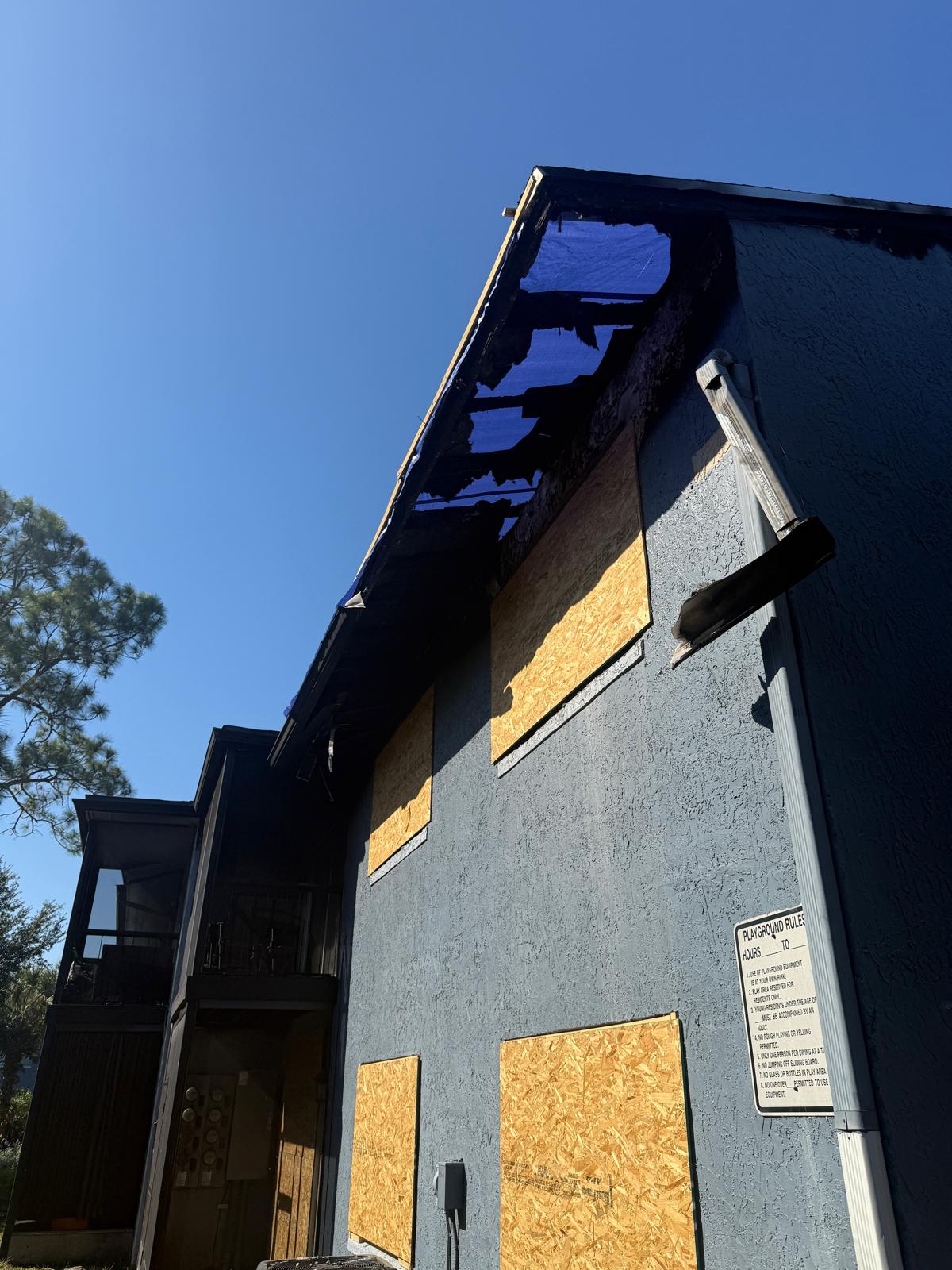 Building with boarded-up windows and roof damage covered by a blue tarp under clear sky.