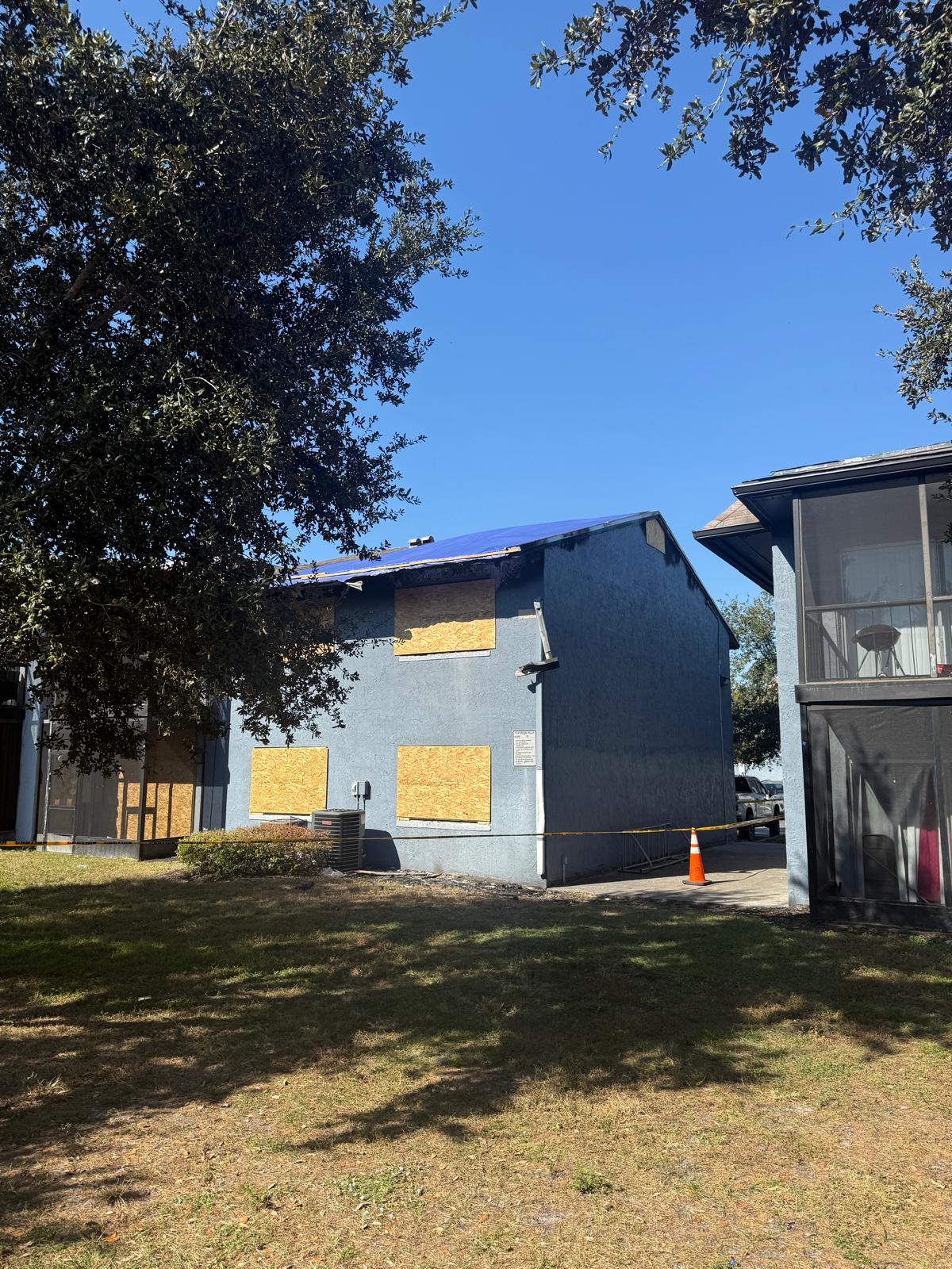 Blue building with boarded-up windows and roof under repair, surrounded by caution tape and an orange traffic cone, with trees and grass in the foreground.