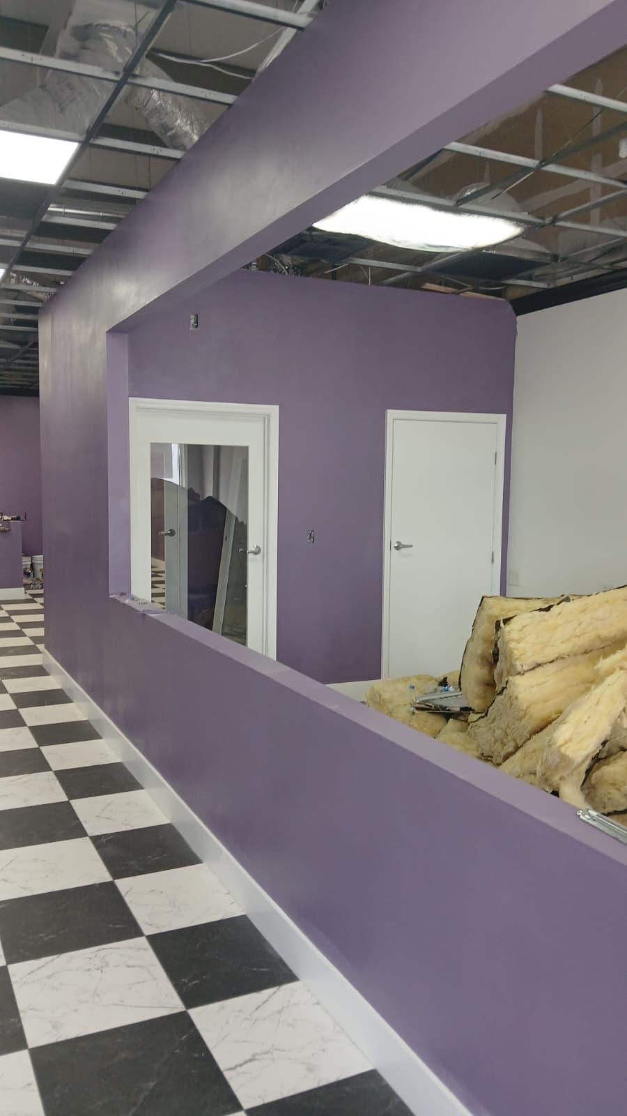Interior hallway with black and white checkered floor, purple walls with a large rectangular window cutout, two white doors, and exposed ceiling with construction insulation.