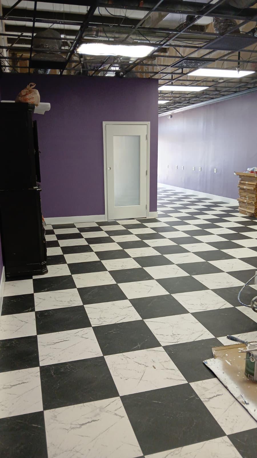 Interior space with black and white checkered floor tiles, purple walls, a white door with a frosted glass panel, and an unfinished ceiling with visible pipes and lights.