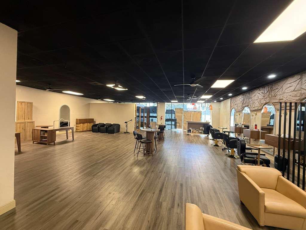 Spacious salon interior with wooden floors, multiple styling stations with chairs and mirrors along right wall, seating area with beige armchairs, and tables with bar stools in the center.